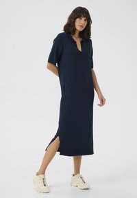 Abito in maglia blu navy con colletto polo, maniche corte e spacchi laterali. Indossato con sneakers crema, che mettono in evidenza una vestibilità rilassata e una trama morbida.