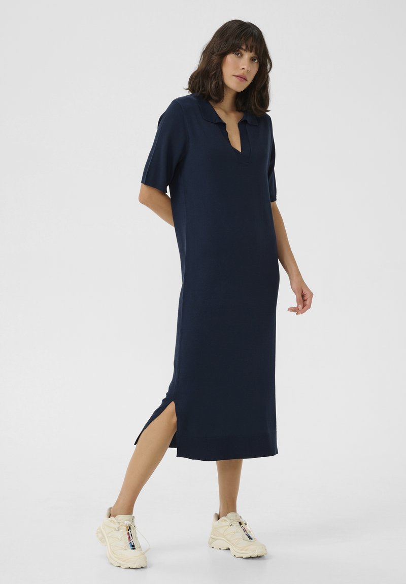 Navy blue knit dress with a polo collar, short sleeves, and side slits. Naisten päällä kermanvalkoiset tennarit, jotka korostavat rentoa istuvuutta ja pehmeää tekstuuria.