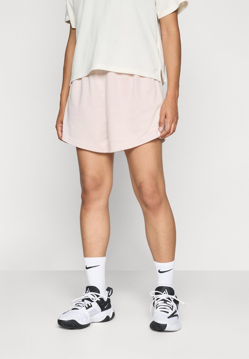 Personne portant des baskets blanches Nike, des chaussettes blanches Nike, un short rose clair et une chemise blanche ample, debout devant un fond uni.