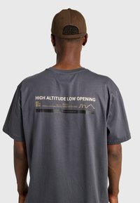 Szara bawełniana koszulka z dużym nadrukiem na plecach zawierającym tekst i grafikę, krótkimi rękawami oraz luźnym krojem.