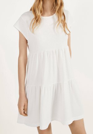 Femme portant une robe blanche à manches courtes et à volants avec un tissu texturé, debout devant un fond clair uni.