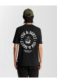 Juodas medvilnės marškinėlis su baltu grafiniu spaudiniu nugarinėje pusėje, įskaitant laivo dizainą ir tekstą "LYLE & SCOTT" su akcentais.
