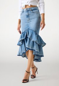 Vrouw in een hoge taille blauwe denim zeemeerminsrok met een gerimpelde zoom en zwarte hoge hakken, staand tegen een lichte achtergrond.