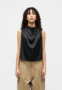 Μαύρο τοπ χωρίς μανίκια με ντραπαρισμένο ντεκολτέ, κατασκευασμένο από γυαλιστερό ύφασμα. Συνδυάζεται με μπεζ, ψηλόμεσα, πλισέ παντελόνια.