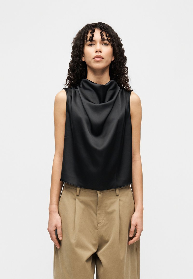 Μαύρο τοπ χωρίς μανίκια με ντραπαρισμένο ντεκολτέ, κατασκευασμένο από γυαλιστερό ύφασμα. Συνδυάζεται με μπεζ, ψηλόμεσα, πλισέ παντελόνια.