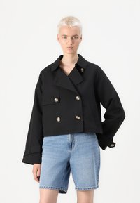 Giacca nera tagliata, a doppiopetto, con colletto a scendere, caratterizzata da bottoni metallici e maniche larghe, abbinata a pantaloni corti in denim azzurro chiaro.