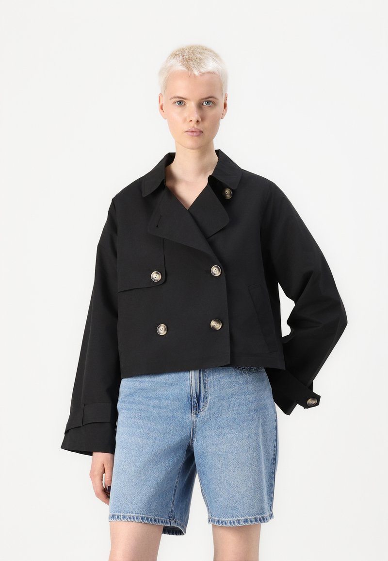 Giacca nera tagliata, a doppiopetto, con colletto a scendere, caratterizzata da bottoni metallici e maniche larghe, abbinata a pantaloni corti in denim azzurro chiaro.
