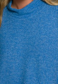 Maglione a maglia blu con texture indossato da una persona con lunghi capelli biondi, visibile dal collo alla parte superiore del petto.
