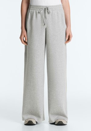 Oseba nosi svetlo sive široke trenirke z elastičnim pasom in vezalko, v kombinaciji z belimi supergami.