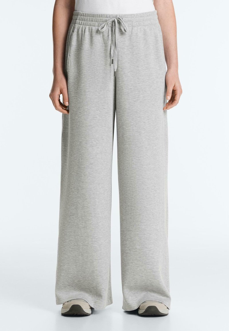 Person iført lysegrå vidde-sweatpants med elastik i taljen og snøre, kombineret med hvide sneakers.