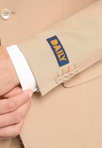 Beżowa rękaw od marynarki z trzema jasnymi guzikami. Ma niebieską metkę z napisem "DAILY" w kolorze pomarańczowym. Częściowo widoczny mankiet białej koszuli pod spodem.