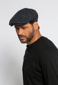 Cappello piatto in lana con motivo a spina di pesce blu scuro, arricchito da sottili macchie bianche. Abbinato a una maglietta henley nera con dettagli di bottoni.