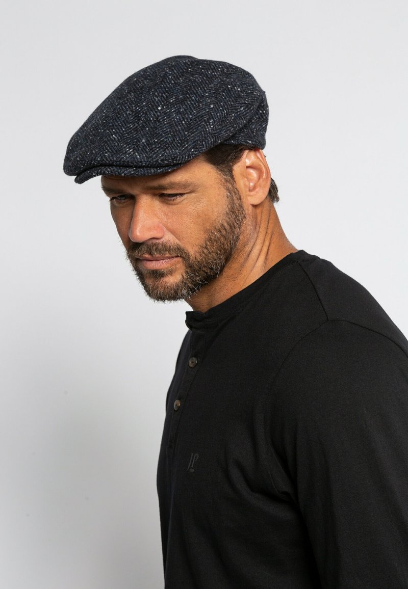 Cappello piatto in lana con motivo a spina di pesce blu scuro, arricchito da sottili macchie bianche. Abbinato a una maglietta henley nera con dettagli di bottoni.