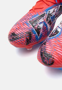 Scarpe da calcio con un motivo testurizzato rosso e blu, con accenti neri e un logo distintivo sulla superficie superiore. Chiodi trasparenti sulla suola.