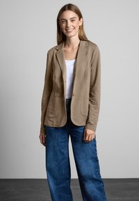 Vaaleanruskea bleiseri, jossa on sileä pinta, viilto-kaulus, kaksi etutaskua, yksittäinen nappikiinnitys, käytetty valkoisen paidan ja sinisten väljien farkkujen kanssa.