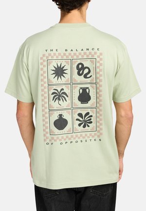 Mężczyzna noszący jasnozieloną koszulkę z sześcioma czarnymi symbolami, w tym słońcem, wężem, palmą i ceramiką oraz napisem „The Balance of Opposites” na plecach.