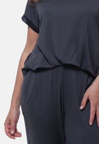 Donkergrijze top met korte mouwen en een losse pasvorm. Beschikt over zachte, gladde stof met een gedrapeerd ontwerp, waardoor zachte plooien rond de taille ontstaan.