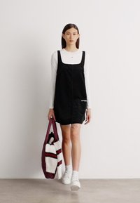 Μαύρη σαλοπέτα με τετράγωνη λαιμόκοψη και πλαϊνές τσέπες, φορεμένη πάνω από λευκή μακρυά μπλούζα με μακριά μανίκια. Το μοντέλο κρατάει μια μπορντό τσάντα ώμου με ρίγες.