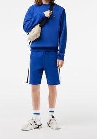 Niebieski zestaw dresowy, składający się z bluzy i szortów, wykonany z miękkiego materiału. Posiada białe paski po bokach, zestawiony z białymi trampkami i beżową torbą na ramię.