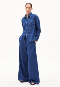 Completo in denim composto da una camicia con colletto e bottoni, dotata di taschino sul petto, e pantaloni a gamba larga. Tutti i capi sono realizzati in un tessuto blu scuro uniforme.
