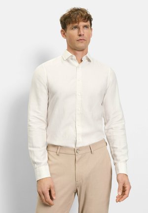 Mann trägt ein langärmeliges weißes Hemd mit Knöpfen und beige Hose, steht vor einem schlichten hellen Hintergrund.