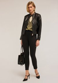 Black leather jacket with a fitted design, gold zipper detail, worn over a green sweater, paired with black skinny jeans and heels, holding a bag.