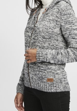 Femme portant un pull en maille gris et blanc à fermeture éclair avec de gros boutons en bois et un col roulé blanc, touchant le pull près de la poitrine.