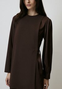 Rochie lungă maro cu mânici lungi și un snur pe lateral. Material neted cu un ușor luciu. Decolteu rotund simplu și umeri structurați, pufoși.