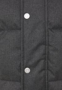 Grå puffervest i fiskebenmønster med quiltet tekstur og blanke sølvknapper. Har en høj krave og vandrette sømdetaljer.