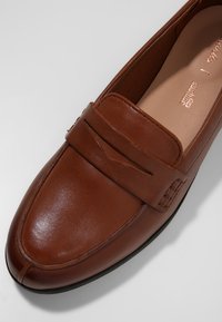 Mocassin en cuir marron avec un design moc toe et une lanière décorative sur le dessus, avec une semelle intérieure beige et une semelle noire.