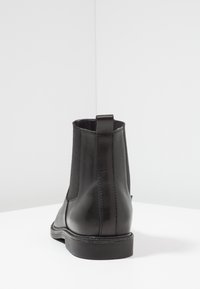 Bottine noire en cuir avec des panneaux élastiques sur les côtés, une sangle à l'arrière et une semelle plate en caoutchouc. Texture lisse et design minimaliste.