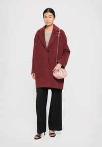 Femme portant un manteau oversize bordeaux, un pantalon large noir, des sandales noires, et tenant un sac rose avec une bandoulière en chaîne.