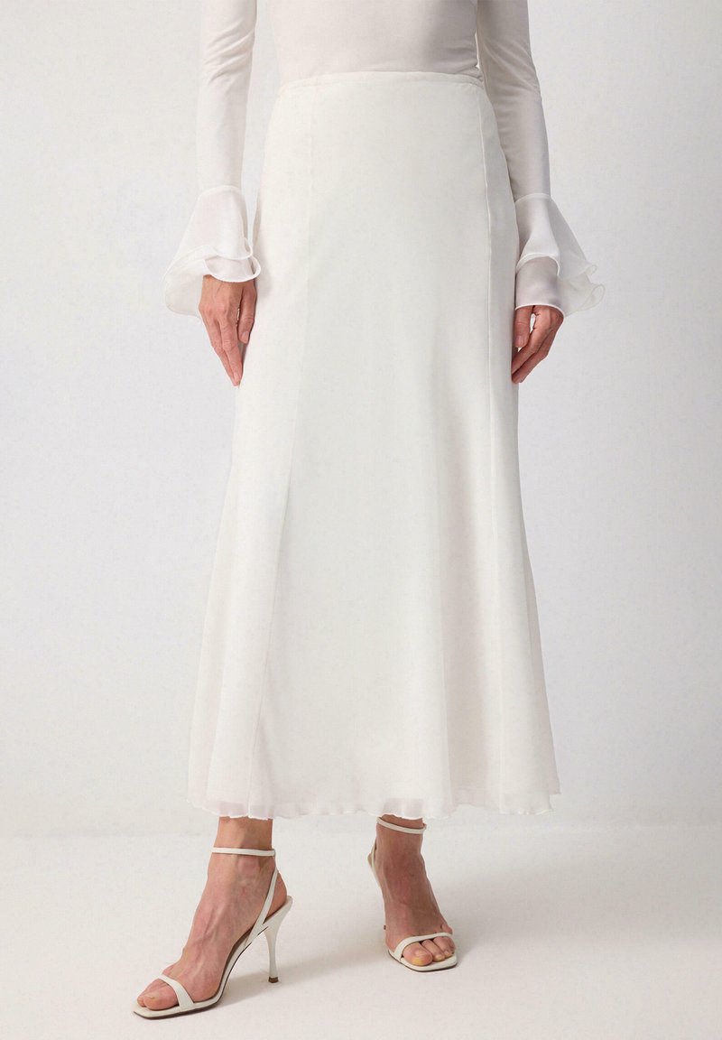 Γυναίκα με λευκή μακριά φούστα, διάφανη μπλούζα με βολάν στα μανίκια και λευκά σανδάλια με ψηλό τακούνι, στέκεται μπροστά σε ένα απλό φόντο.