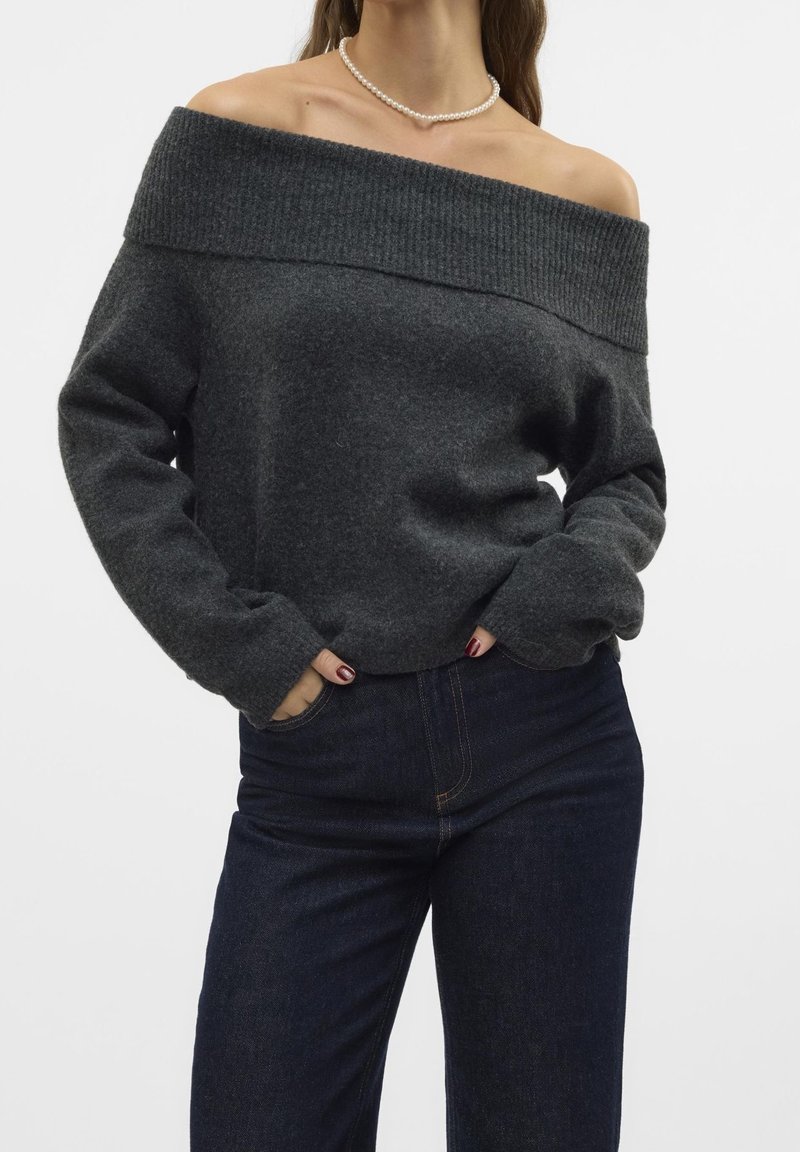 Femme portant un pull gris foncé à épaules dénudées, un jean bleu foncé, un collier de perles et un vernis à ongles rouge foncé, mains dans les poches.