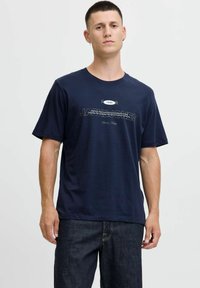 Tamsiai mėlynas medvilninis marškinėlis trumpomis rankovėmis, apvaliu iškirpimu ir priekyje esanti grafika su tekstu "JACK & JONES 1990" kontrastingomis baltomis ir juodomis spalvomis.