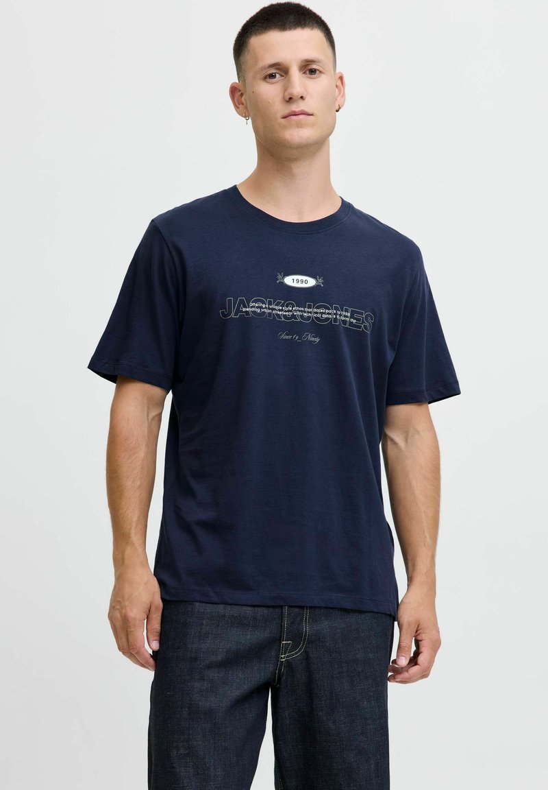 Tamsiai mėlynas medvilninis marškinėlis trumpomis rankovėmis, apvaliu iškirpimu ir priekyje esanti grafika su tekstu "JACK & JONES 1990" kontrastingomis baltomis ir juodomis spalvomis.