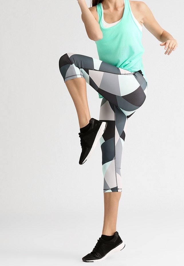 Personne en leggings à motif géométrique et débardeur menthe levant un genou en faisant de l'exercice sur un fond uni.