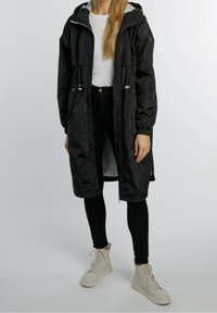 Schwarzer langer Mantel mit Kordelzugkapuze, zwei Seitentaschen und elastischen Bündchen über einem weißen Oberteil und schwarzen Hosen, kombiniert mit weißen High-Top-Sneakers.