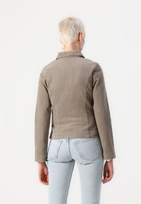 Osoba o krótkich blond włosach, nosząca srebrne kolczyki-koła, taupe'ową kurtkę sztruksową i jasnoniebieskie jeansy, zwrócona tyłem na białym tle.