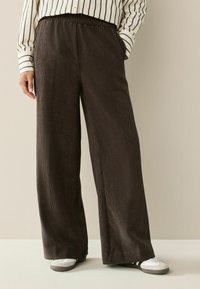 Weite, dunkelbraune Cordhosen mit strukturierter Oberfläche, elastischem Bund und seitlichen Taschen, kombiniert mit weißem Schuhwerk.