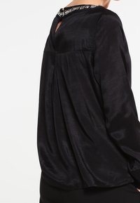 Blouse en soie noire à coupe ample, dos froncé et détail orné autour de l'encolure. Comprend des manches longues et une ouverture en forme de goutte au dos.