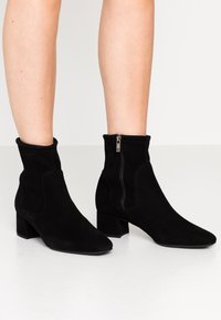 Gambe che indossano stivaletti neri in suede con tacco a blocco e cerniere laterali, in piedi su un pavimento bianco contro uno sfondo chiaro e semplice.