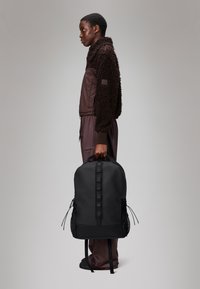 Mochila preta com um acabamento suave, alças ajustáveis, bolsos laterais em malha e uma marcação discreta. Modelo vestindo uma jaqueta de fleece castanha.