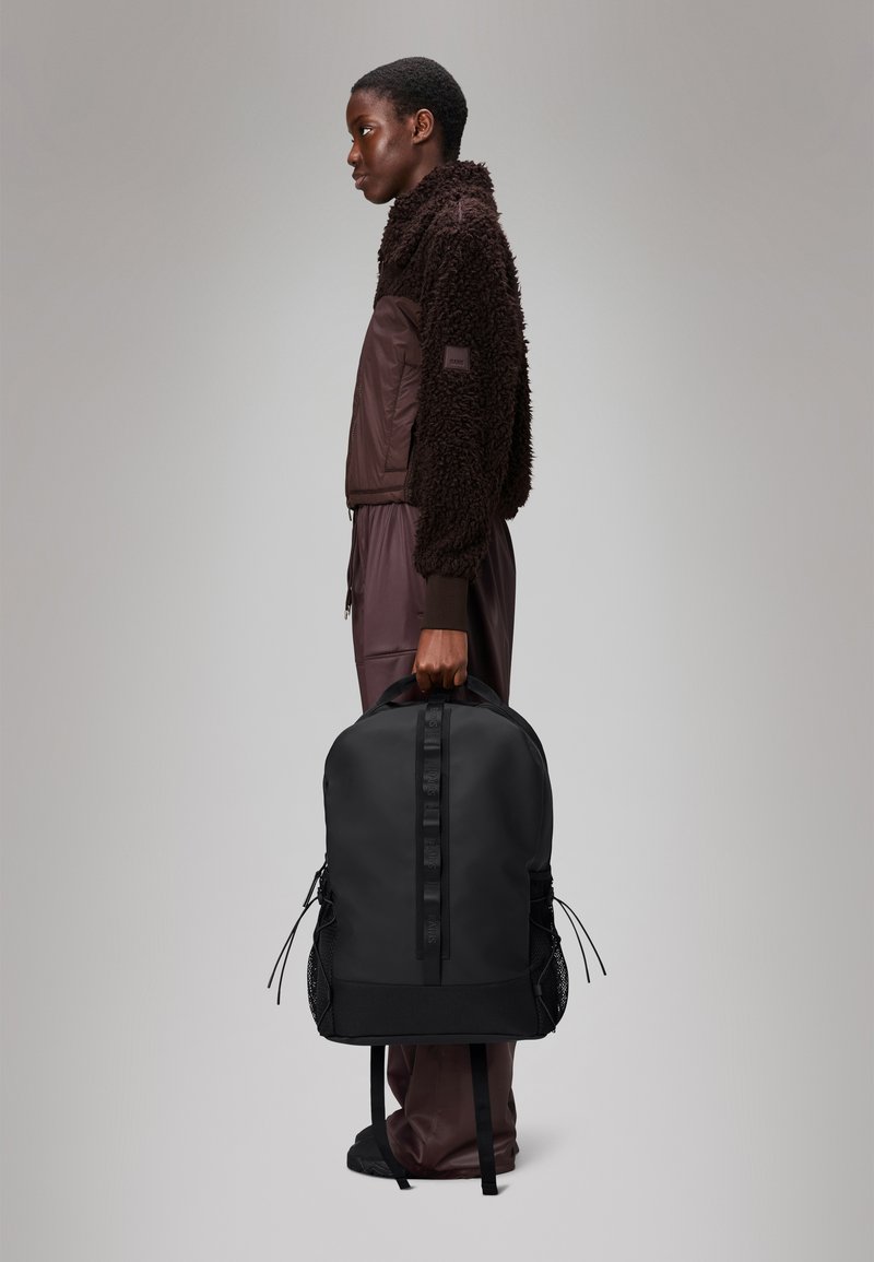 Mochila preta com um acabamento suave, alças ajustáveis, bolsos laterais em malha e uma marcação discreta. Modelo vestindo uma jaqueta de fleece castanha.
