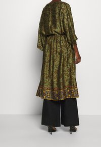 Zielona sukienka w wzory z szerokimi rękawami, elastycznym pasem i kontrastowym wykończeniem hemu. Połączona z szerokimi czarnymi spodniami i butami na obcasie.