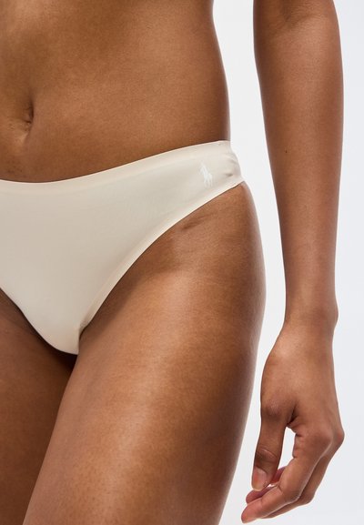 Γυναίκα που φοράει μπεζ εσώρουχα μπικίνι με μικρό λευκό λογότυπο στο αριστερό ισχίο, με το χέρι χαλαρό δίπλα της, πάνω σε απλό φόντο.