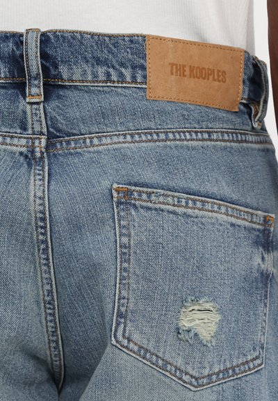 Gai zila denim bikses ar nolietotu aizmugures kabatu, kas aprīkota ar brūnu ādas plāksni, uz kuras ir uzraksts "THE KOOPLES" jostas vietā.