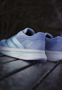 Helle blaue Laufschuhe mit strukturiertem Mesh-Obermaterial, drei kontrastierenden Streifen und einer weißen Zwischensohle mit einem hellgrünen Akzent.