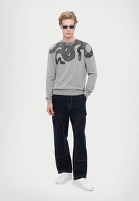 Felpa grigia con grafica nera di un serpente sulla spalla, abbinata a pantaloni blu navy con dettagli di cucitura rossa e sneakers bianche.