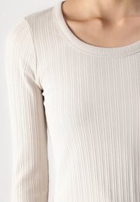Ribbed long-sleeve top in light beige, featuring a round neckline and soft texture, emphasizing a fitted silhouette.